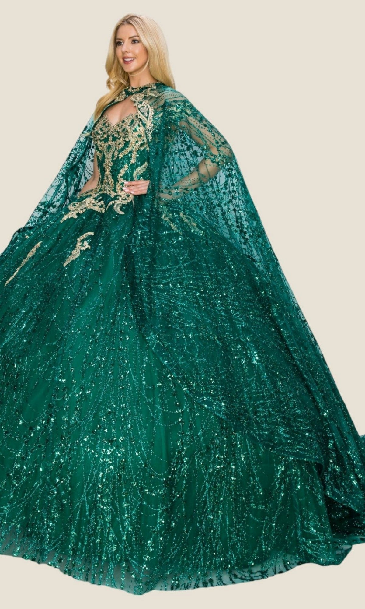 A woman wearing the Quinceanera Dress 4201 By Cinderella Couture—a voluminous emerald green ball gown with gold embroidery and a matching sheer cape—stands against a neutral background.